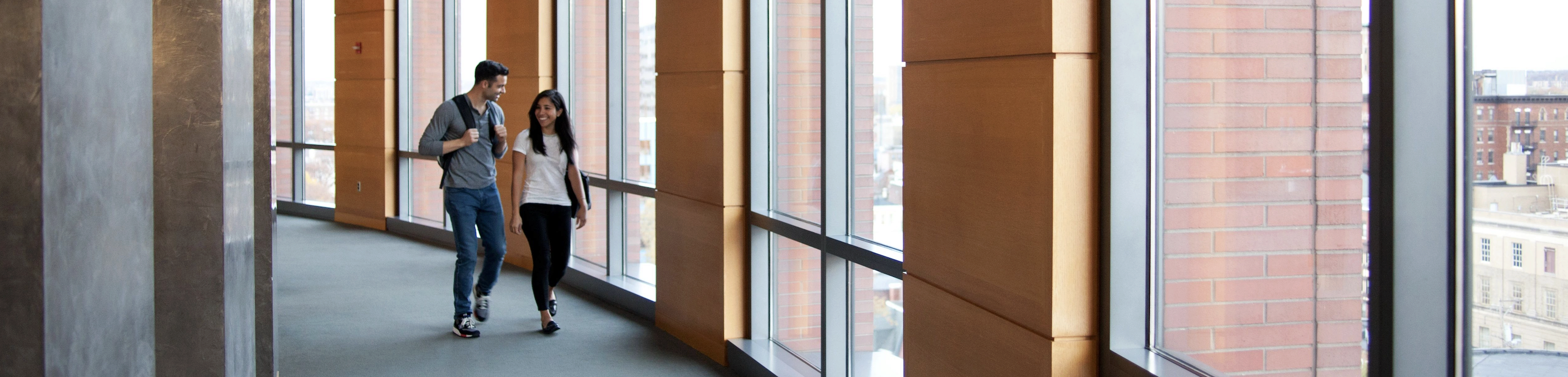 A couple of people walking through a bright, modern hallway with large windows and wooden paneling.